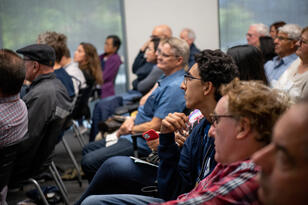 Several people sit in chairs in a lecture hall listening.